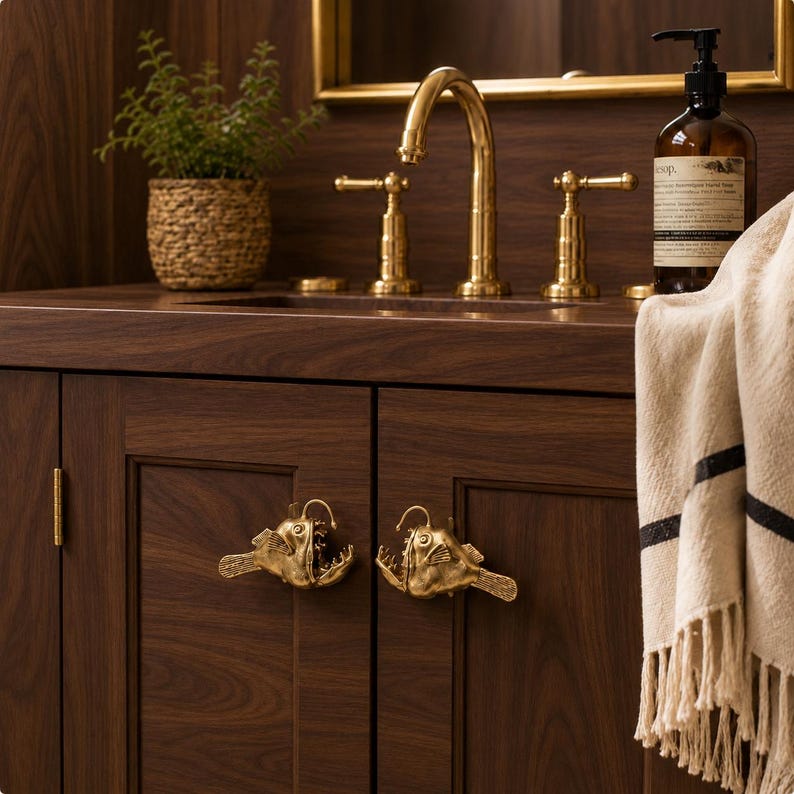 May include: Two brass cabinet knobs shaped like anglerfish with detailed fins and open mouths, mounted on dark wood cabinet doors. The knobs feature a metallic gold finish and are positioned horizontally on the front of the vanity doors.