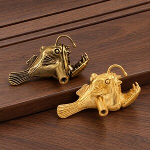 May include: Two anglerfish-shaped cabinet knobs, one in an antique brass finish and one in a bright gold finish. Each knob features a detailed fish body with a prominent lure, sharp teeth, and a cylindrical mounting post with a threaded hole.