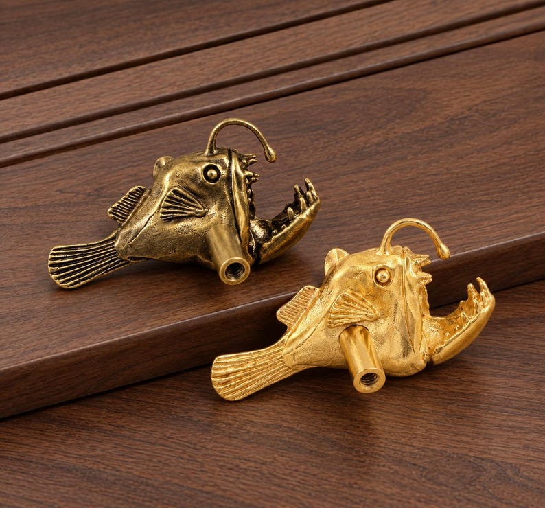 May include: Two anglerfish-shaped cabinet knobs, one in an antique brass finish and one in a bright gold finish. Each knob features a detailed fish body with a prominent lure, sharp teeth, and a cylindrical mounting post with a threaded hole.