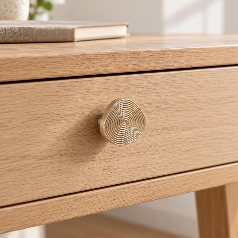 May include: A gold-toned metal drawer knob with an organic, irregular shape and a textured surface featuring concentric, swirling circular lines. The knob is mounted on the front of a light-coloured wooden drawer with a visible grain pattern.