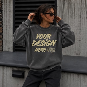 May include: A charcoal grey crewneck sweatshirt with a vintage-style wash, featuring the beige text "YOUR DESIGN HERE" printed in a bold, brush-stroke font across the chest, accompanied by the smaller script text "MOCKUP ATELIER" below it.