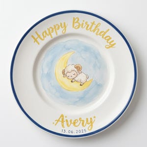 Puede incluir: Un plato de cerámica blanca con borde azul oscuro que presenta una ilustración en acuarela de una oveja durmiendo sobre una luna creciente amarilla sobre una nube azul. El texto dice Happy Birthday en oro, y Avery 13.06.2025 en oro y azul oscuro.