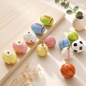 May include: A collection of ceramic cabinet knobs shaped like various sports balls, including baseballs, footballs, footballs, and basketballs. The knobs feature colourful finishes with painted red stitching or black patterns, each equipped with a metal screw.
