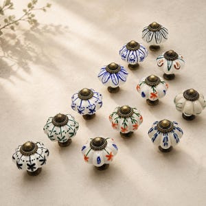 Könnte beinhalten: Eine Sammlung von zwölf runden, geriffelten Keramik-Schrankknöpfen mit verschiedenen handbemalten Blumen- und geometrischen Mustern in Blau, Grün, Orange und Schwarz. Jeder Knopf ist mit einem kleinen, runden Messingbeschlag versehen.