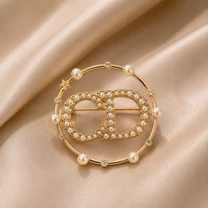 May include: A gold-toned circular brooch featuring the letters CD in the center, embellished with small white faux pearls. The outer ring is accented with alternating faux pearls and clear crystals, with a small star detail on the upper left side.
