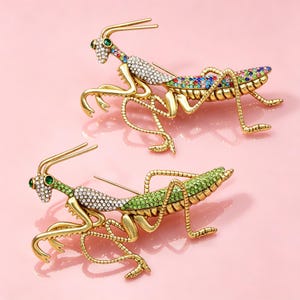 May include: Two gold-toned metal praying mantis brooches. The top brooch features multicolored crystals on the body and clear crystals on the thorax. The bottom brooch features green crystals on the body and clear crystals on the thorax. Both have green eyes.
