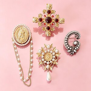 May include: A collection of four gold-tone metal brooches. One is a cross with red stones, one is an oval coin brooch with dangling pearl chains, one is a floral-shaped brooch with a central coin and pearl drop, and one is a C-shaped pearl brooch.