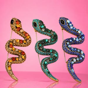 Puede incluir: Tres broches con forma de serpiente y cuerpos de metal dorado incrustados con pequeños diamantes de imitación. Cada broche muestra una paleta de colores distinta: uno con piedras ámbar y naranja, otro verde esmeralda y otro con cristales azules e iridiscentes.