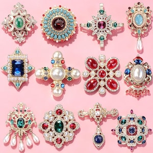 May include: A collection of twelve ornate metal brooches featuring various designs with embedded faux pearls, colorful rhinestones, and crystals in shades of red, blue, green, and clear, arranged in a grid on a solid light pink background.