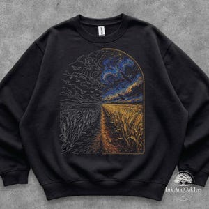 May include: A black crewneck sweatshirt featuring a split-design graphic. The left side shows a white line-art illustration of a wheat field and birds, while the right side displays a colorful, detailed painting of a wheat field under a starry sky.