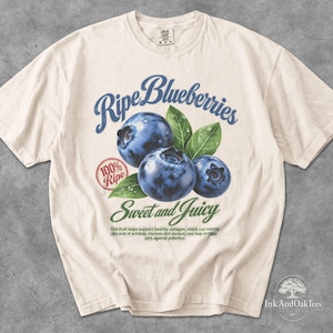 May include: A cream-coloured short-sleeve t-shirt featuring a graphic of three blueberries with green leaves. The text reads Ripe Blueberries, 100% Ripe, Sweet and Juicy, and a small paragraph about fruit benefits. The shirt has a crew neckline.