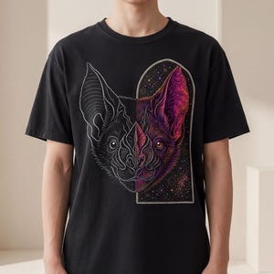 May include: A black short-sleeve t-shirt featuring a split-design graphic of a bat head. The left side is a white line drawing, while the right side is a colorful, detailed illustration of a bat set within an arched frame filled with stars.