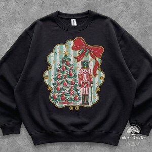 Puede incluir: Una sudadera negra de cuello redondo con un gráfico central de un árbol de Navidad decorado y un soldado cascanueces. El diseño está enmarcado por un borde festoneado con rayas azul claro, detalles dorados y un gran lazo rojo.