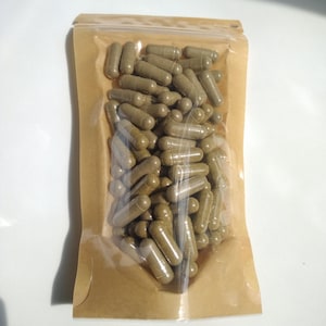 Pode incluir: Uma bolsa de plástico transparente com verso de papel castanho cheia de numerosas cápsulas cilíndricas verde-azeitona. A bolsa possui um fecho selável e contém uma grande quantidade de cápsulas idênticas.