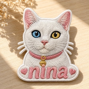 Op de afbeelding: Een geborduurde patch met een witte kat met één blauw oog en één geel oog. De kat draagt een roze halsband met een gouden belletje. Onder de kat staat de naam nina geborduurd in roze letters, geflankeerd door twee kleine roze hartjes.