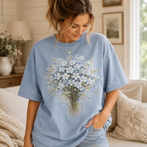 May include: A light blue short-sleeve t-shirt featuring a large, detailed graphic print of a bouquet of light blue forget-me-not flowers with green stems and leaves centered on the front. The shirt has a crew neckline and a relaxed, loose fit.