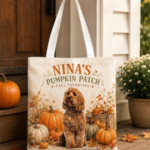 May include: A cream-colored canvas tote bag with white handles featuring a printed graphic of a brown curly-haired dog sitting among pumpkins and autumn leaves. The text reads NINA'S PUMPKIN PATCH FALL FAVORITES in brown and green lettering.