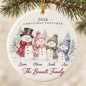 Può includere: Un addobbo natalizio rotondo in ceramica bianca con un'illustrazione ad acquerello di quattro pupazzi di neve con cappelli e sciarpe. Il testo recita 2026 CHRISTMAS TOGETHER, i nomi Liam, Olivia, Noah, Ava e The Bennett Family in corsivo rosso.