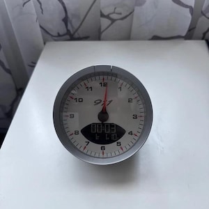 May include: A round analogue clock with a silver bezel and a white face featuring black numerals from 1 to 12. The centre displays the text 911 in a stylised font with a red needle. A small digital screen at the bottom shows the time 00:03.