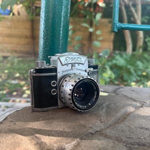 Könnte beinhalten: Eine Vintage-Kamera vom Typ Exa von Ihagee Dresden mit silbernem Metallgehäuse und schwarzem, texturiertem Griff. Die Vorderseite zeigt einen silbernen Objektivtubus mit der Gravur Meyer-Optik Görlitz Primotar 2.8/50 und der Nummer 2240431.