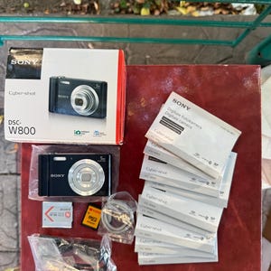 May include: A black Sony Cyber-shot DSC-W800 digital camera with its original white and red box, a rechargeable battery, a memory card adapter, a charging cable, and a stack of multiple instruction manuals in various languages.