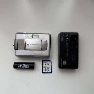 May include: A silver Konica Minolta Dimage X50 digital camera, a black BC-800 lithium-ion battery charger, a black eFilm battery, and a blue and white Canon 16MB SD memory card. The camera features a sliding lens cover and a built-in flash.