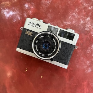 May include: A silver and black Minolta Hi-Matic F 35mm film camera. The front features a lens with text reading Minolta Camera Co. Ltd. Rokkor 1:2.7 f=38mm. The top plate displays the Minolta Hi-Matic F logo, and a small square emblem is on the left.
