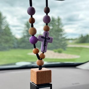 Pode incluir: Um aromatizador de carro pendurado no espelho retrovisor, com um frasco de vidro quadrado preto e tampa de madeira, rotulado Teakwood Mahogany. Um cordão preto sustenta contas de madeira roxas e castanhas e uma cruz roxa com a palavra Jesus.