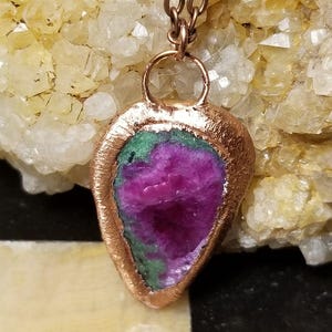 Puede incluir: Un colgante en forma de lágrima que presenta un cristal de rubí magenta crudo y vibrante rodeado de zoisita verde, engastado en un marco de cobre metálico texturizado. El colgante cuelga de una cadena de eslabones de metal color cobre.