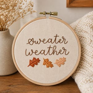 May include: A circular wooden embroidery hoop containing off-white fabric with the brown stitched text "sweater weather" and three embroidered oak leaves in varying shades of orange, rust, and tan positioned below the text.