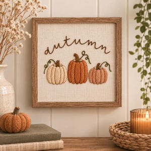 Könnte beinhalten: Ein quadratischer Holzrahmen mit einem cremefarbenen Stoff und brauner Stickerei. Die Stickerei zeigt das Wort autumn in Schreibschrift über drei Kürbissen in den Farben Creme, Orange und gedecktem Rosa mit grünen Ranken.