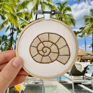 May include: A circular wooden embroidery hoop containing white fabric with a hand-stitched design of a spiral seashell. The shell is rendered in light beige thread with dark brown outlines, featuring segmented chambers that follow the spiral pattern.