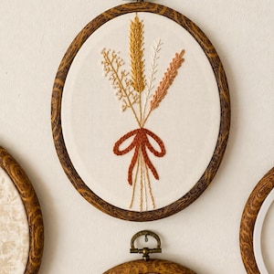 Op de afbeelding: Een ovale borduurring met een crèmekleurige stoffen achtergrond en een handgestikt ontwerp van een boeket tarwe en wilde bloemen, samengebonden met een roestkleurige strik. De lijst heeft een donkerbruine houtnerf en een koperen ophanghaak.