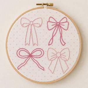May include: A circular wooden embroidery hoop containing white fabric with a light pink polka dot pattern. Four distinct bows are embroidered on the fabric using various shades of pink thread, featuring different styles and knot configurations.