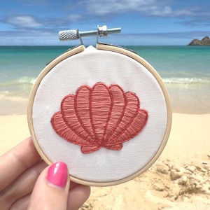 May include: A circular wooden embroidery hoop containing white fabric with a coral-colored thread design of a seashell. The hoop features a metal tension screw at the top, securing the fabric in place for the detailed stitched shell illustration.