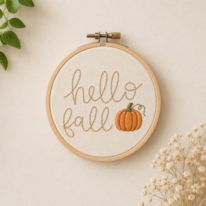 Può includere: Un telaio da ricamo circolare in legno contenente un tessuto bianco sporco con il testo "hello fall" ricamato a mano in filo marrone chiaro, accompagnato da una piccola zucca arancione ricamata con un gambo verde e un dettaglio a viticcio.