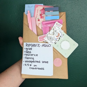 May include: A brown paper-wrapped book package featuring a white note labeled ROMANCE-ADULT with a list of themes including grief, loss, resilience, family, and unexpected love. Accessories include a pink pen, blue page markers, and stickers.