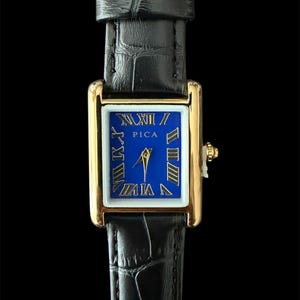 Könnte beinhalten: Eine rechteckige Armbanduhr mit goldfarbenem Gehäuse und einem leuchtend blauen Zifferblatt mit goldenen römischen Ziffern. Auf dem Zifferblatt steht der Schriftzug PICA in Gold. Sie hat ein schwarzes Lederarmband mit strukturierter Oberfläche.