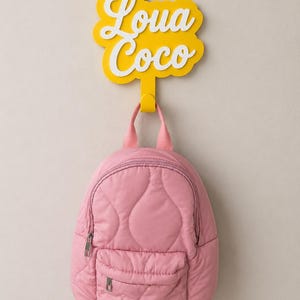 Op de afbeelding: Een gele wandhaak met een wolkvormig bordje met de witte tekst Loua Coco in een schrijflettertype. Een roze gewatteerde rugzak met een voorvak met rits en zilverkleurige details hangt aan de haak en vertoont een zachte textuur.