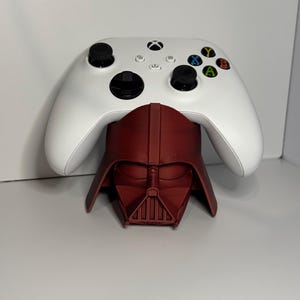 May include: A white wireless video game controller resting on a dark red, 3D-printed stand shaped like the helmet of Darth Vader. The controller features black thumbsticks, a black directional pad, and multicolored action buttons labeled A, B, X, and Y.