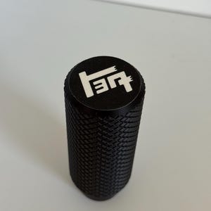 May include: A cylindrical black metal gear shift knob featuring a textured diamond-patterned grip along its side. The top surface is flat and black, displaying a white engraved logo that resembles the letters T and E arranged in a stylized geometric design.