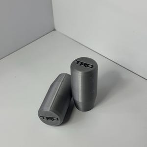 May include: Two cylindrical gear shift knobs in a matte grey finish. Each knob features a tapered base and a flat top surface with the letters TRD embossed in black. The surface texture shows subtle horizontal lines from the manufacturing process.