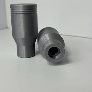 May include: Two cylindrical gray objects with a smooth finish. One object stands upright, featuring a flat top and horizontal grooves near the upper edge. The second object lies on its side, revealing a hollow circular opening at the center.