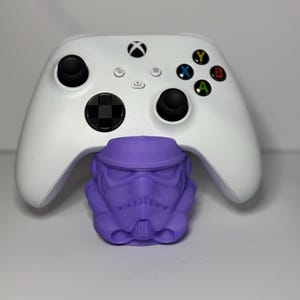 May include: A white wireless video game controller with black analog sticks and a D-pad, positioned behind a purple 3D-printed stormtrooper helmet stand. The controller features the Xbox logo and colored action buttons in blue, yellow, green, and red.