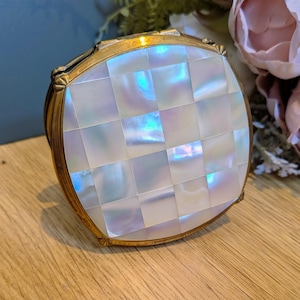 May include: A round compact case featuring a gold-toned metal frame and a lid inlaid with a grid of iridescent, square mother-of-pearl tiles. The surface of the tiles displays a shimmering, pearlescent finish with subtle blue and pink highlights.
