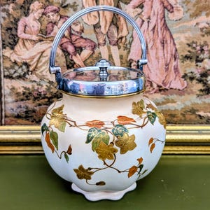 May include: A cream-coloured ceramic biscuit barrel with a scalloped base and a silver-toned metal lid featuring a swing handle. The body is decorated with a repeating pattern of gold, green, and orange leaves and flowers on a vine motif.