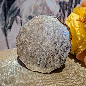 May include: A round, silver-toned metal compact mirror featuring an intricate, embossed floral and scrollwork pattern across its entire surface. The edges of the compact are scalloped, and the metal has a textured, matte finish with polished highlights.