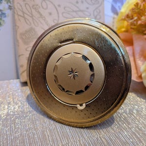 May include: A round gold-tone metal powder compact featuring a textured surface with a central circular medallion. The medallion has a scalloped edge and a small engraved star design in the centre. The compact has a hinged lid and a small latch.