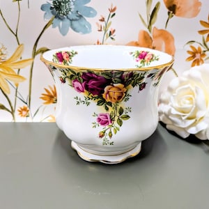 May include: A white porcelain bowl with a scalloped rim and gold trim, featuring a floral pattern of pink, yellow, and deep red roses. The bowl has a curved, flared shape with a decorative base, showcasing detailed botanical illustrations.