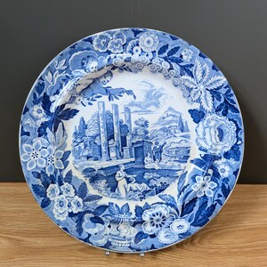 May include: A round ceramic plate featuring a blue and white transferware pattern. The centre depicts a classical landscape with ruins and figures, while the wide rim is decorated with detailed floral motifs and foliage in shades of blue.